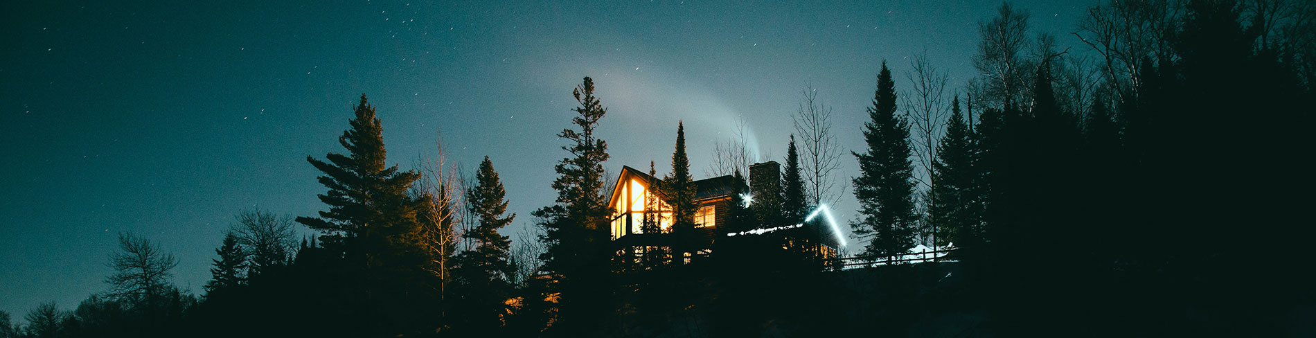 A cottage sits on a ridge, lit up at night with trees to either side and the night sky