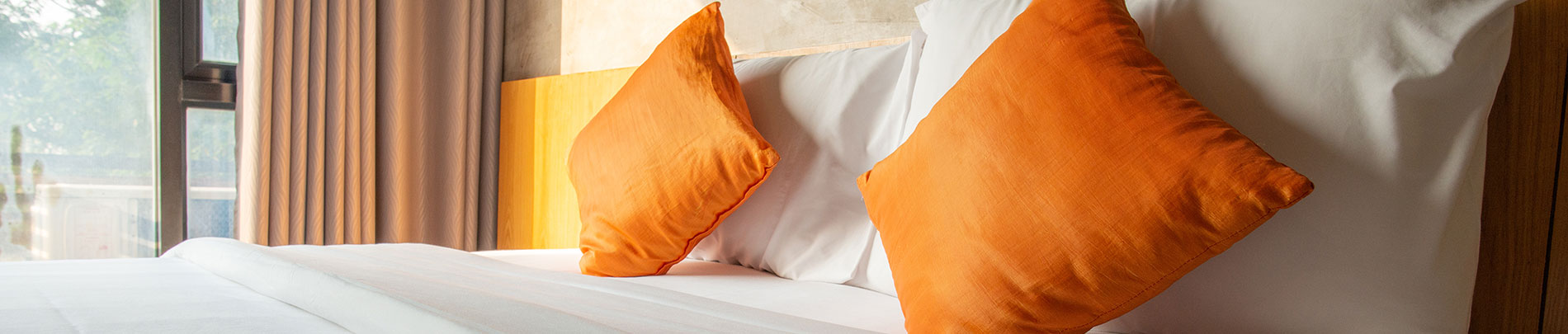 Bright coloured pillows rest on a freshly made bed with light streaming in from a glass door.